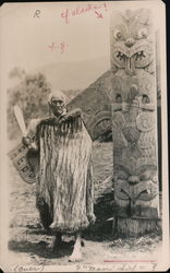 Rewi Manga Maniapoto is wearing a Korowai traditional cloak, holding a greenstone club (mere pounamu) and standing in front of a carved meeting house (wharenui). Postcard