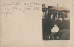 A young child wearing a white gown is sitting in a pull wagon that has wire spoke wheels. Postcard