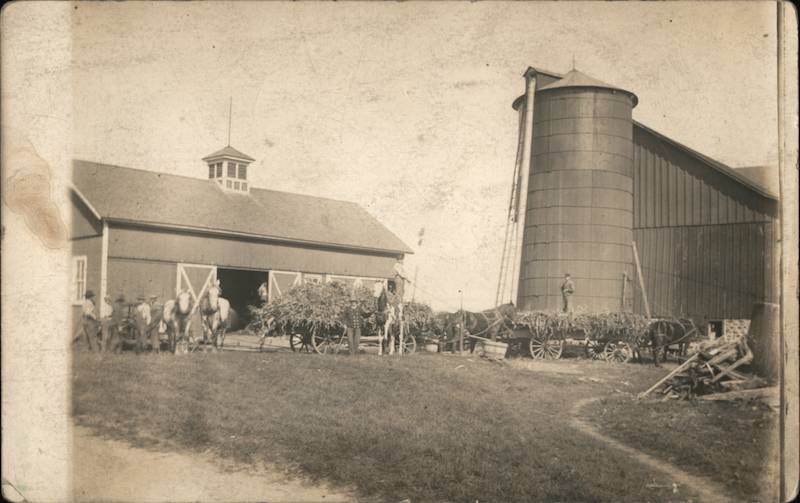 Farm with silo. Several farmers stand in front with cows and wagons of hay.