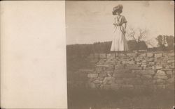 a Well Dressed Woman with a hat Standing atop a Tall Rock Wall Postcard