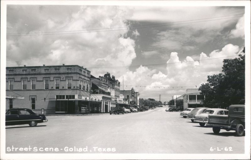 Street scene with vintage cars, storefronts, and cloudy sky Goliad Texas