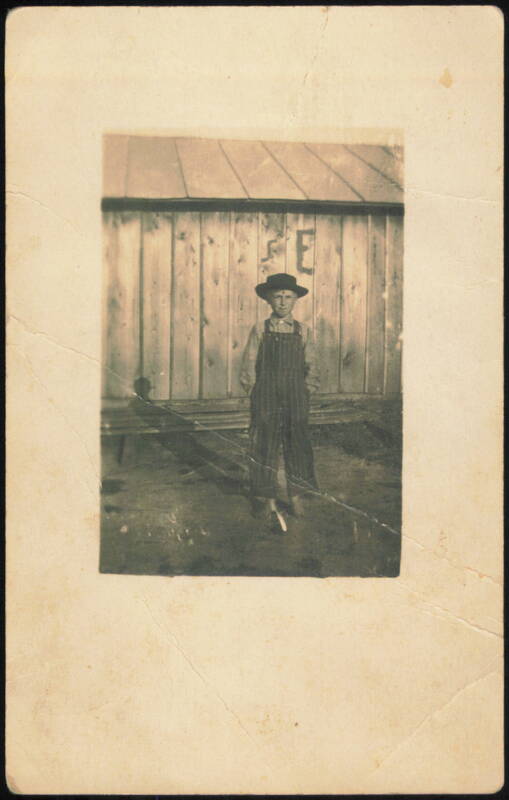 Boy in Overalls and Hat by Wooden Building Children