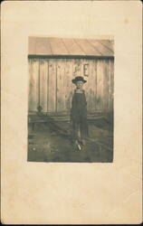 Boy in Overalls and Hat by Wooden Building Postcard