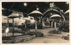 A Fountan and Large Mushrooms Surrounded by Landscaping and an Archway Postcard