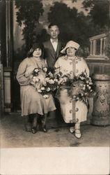 A Man Standing Behind Two Women Holding Flowers Postcard
