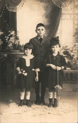 Three children dressed up, posing with flowers and parasol Postcard