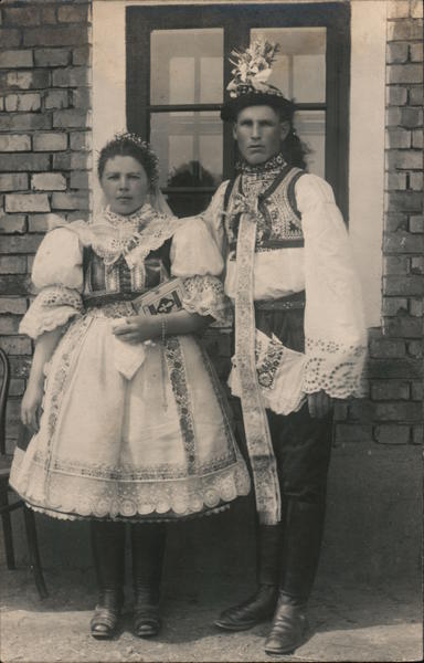 Man and Woman in native costume pose by a brick building Postcard