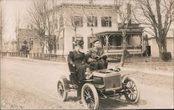 Man and woman driving an early automobile past houses on a dirt road Postcard