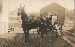 A man and woman riding in a horse and buggy in front of a barn Postcard