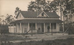 Man and woman pose on large patio of their home in a large clearing Postcard