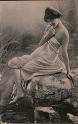 Young woman wrapped in a white cloth is reclining on a rock over a river Postcard